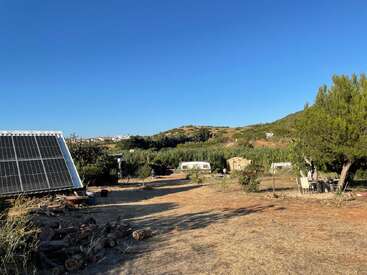 Uma paisagem rural apresenta painéis solares, caravanas, árvores e um campo seco e gramado sob um céu azul claro. Colinas e vegetação dispersa preenchem o plano de fundo.