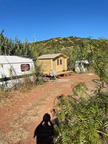 Uma pequena cabana de madeira e duas caravanas estão localizadas em uma área rural com solo seco, grama e arbustos. Atrás, uma colina e um céu azul claro.