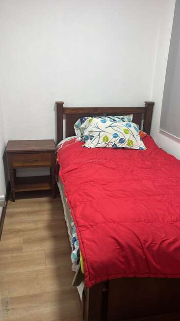 Ein kleines Schlafzimmer mit einem Einzelbett aus Holz, einer roten Bettdecke, einem gemusterten Kissen, einem Nachttisch aus Holz, weißen Wänden und einem Holzboden, das eine klare, gemütliche Atmosphäre schafft.
