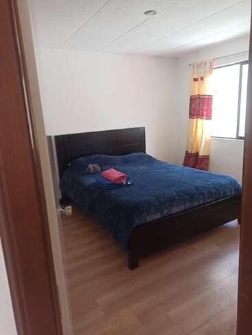 Dieses Bild zeigt ein schlichtes, aufgeräumtes Schlafzimmer mit Holzboden. Zu sehen sind ein Bett mit blauem Bettbezug, gefaltete Handtücher, ein Fenster und hereinströmendes Sonnenlicht.