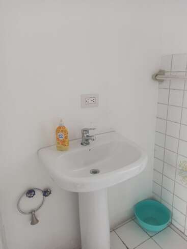 Ein kleines weißes Waschbecken mit einem einzigen Wasserhahn, Flüssigseife, einem Handtuchring, einer Steckdose darüber, einer gefliesten Wand daneben und einer blauen Plastikschüssel darunter. Eine schlichte Badezimmerausstattung.