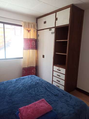 Ein gemütliches Schlafzimmer mit einer blauen Decke, gefalteten Handtüchern, einem großen Fenster mit gelben Vorhängen und einem Kleiderschrank aus Holz mit weißen Türen und offenen Regalen. Helle Beleuchtung.
