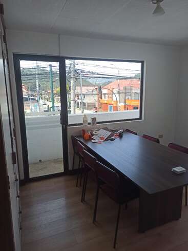 Das Bild zeigt einen kleinen Besprechungsraum mit einem rechteckigen Holztisch, acht Stühlen, herumliegenden Papieren, einem großen Fenster und einem Blick auf die Straße draußen.