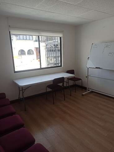Das Bild zeigt einen schlichten, hellen Raum mit einem Fenster, einem weißen Tisch, Stühlen, einer Whiteboard-Tafel mit Zeichnungen und einem Holzboden.