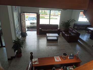 Das Bild zeigt eine gemütliche Hotel- oder Bürolobby mit braunen Ledersofas, einem Couchtisch, Topfpflanzen, einer Rezeption, einer offenen Tür und einem draußen geparkten Auto.