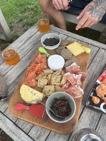 A wooden table holds a charcuterie board with cheeses, crackers, prosciutto, salami, spreads, and two drinks. Two people sit outdoors enjoying the picnic.