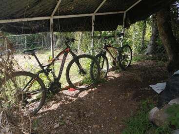 Two mountain bikes are parked under a shaded structure with a wire fence surrounding them. Sunlight filters through trees and foliage, casting shadows on the ground.