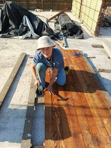 A person wearing a hat works outdoors on wooden planks, kneeling and using a tool. Construction materials and tarps are scattered in the background. Sunlight shines brightly.