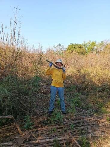 A person stands outdoors in a field, wearing a yellow shirt, blue gloves, jeans, and a hat, smiling and holding a machete over their shoulder.