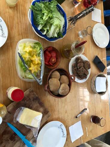 A wooden table set for a meal with plates, lettuce, bread rolls, hamburger patties, sliced tomatoes, cheese, mayonnaise, and beverages, ready for assembling sandwiches.