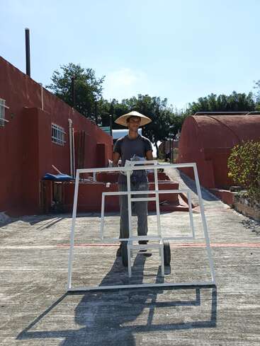 Una persona con un sombrero cónico está de pie al aire libre en un día soleado, sosteniendo una estructura metálica blanca con ruedas, rodeada de edificios rojos y vegetación.