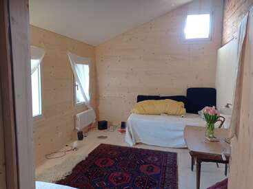 Esta acogedora habitación tiene paredes de madera clara, un sofá blanco con cojines amarillos, una alfombra roja estampada, ventanas con cortinas, una mesa y flores rosas.