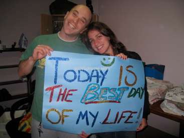 The image depicts a man and woman holding a sign that reads "Today is the best day of my life!" in a room with various items.