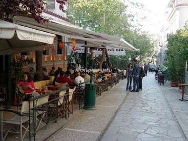 L'image représente un café en plein air, avec une rangée de tables et de chaises, et quelques personnes debout à proximité, avec des arbres et des bâtiments en arrière-plan.