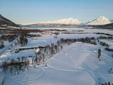 La imagen representa un sereno paisaje nevado con un edificio, árboles y montañas de fondo, mostrando una apacible escena invernal.