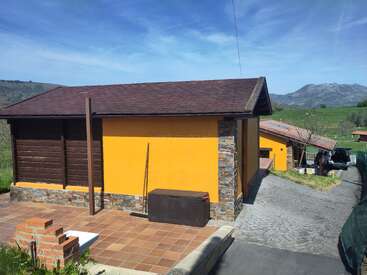 Uma casa amarela com telhado marrom fica em um pátio de ladrilhos, com detalhes em pedra. Um depósito externo, montanhas e campos verdes são visíveis sob um céu azul.