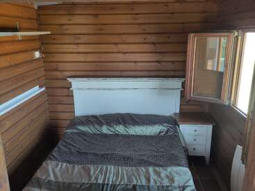 Este aconchegante quarto de madeira tem uma cama de casal, cabeceira branca, duas mesinhas de cabeceira brancas, uma janela aberta, prateleiras e luz natural entrando no espaço compacto.
