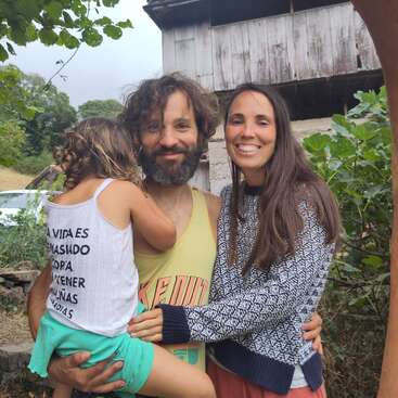 Uma família feliz de três pessoas posa ao ar livre, rodeada de vegetação. O homem segura uma menina nos braços, enquanto uma mulher os abraça; todos sorriem calorosamente para a câmera.