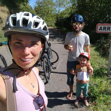 A family of three wearing bike helmets smiles outdoors, standing near bikes and a sign. The child and adult playfully stick out their tongues, enjoying a sunny day.
