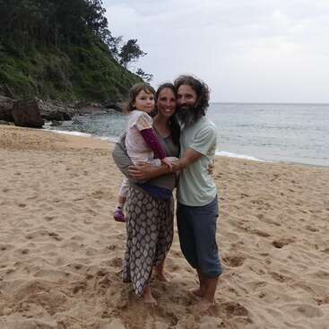 A joyful family of three stands on a sandy beach, smiling and embracing. The ocean and a green, tree-covered hillside provide a beautiful, peaceful backdrop.