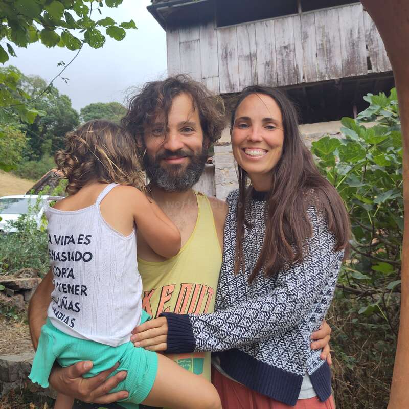 Uma família feliz de três pessoas posa ao ar livre, rodeada de vegetação. O homem segura uma menina nos braços, enquanto uma mulher os abraça; todos sorriem calorosamente para a câmera.