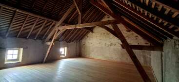 This image shows a spacious, empty attic room with wooden beams, slanted ceiling, old stone walls, two small windows, and a wooden floor, illuminated by natural light.
