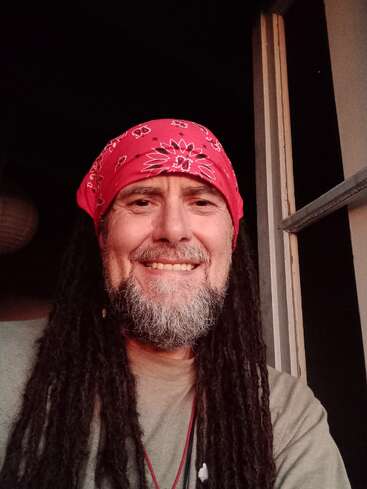 A smiling man with long dreadlocks and a gray beard wears a red bandana. He is indoors, possibly near a window, with warm lighting on his face.