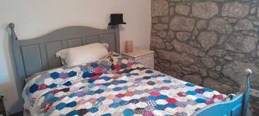 Une chambre confortable avec un lit en bois gris, une couette patchwork colorée, un oreiller simple, un mur en pierre, une table de chevet, une lampe tissée et un chapeau noir.