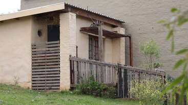 This image shows a small, rustic building with a sloped roof, wooden fence, overgrown plants, and a simple porch. The surrounding area is grassy and green.