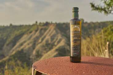 A bottle of olive oil stands on a rusty surface outdoors. In the background, scenic hills and greenery under a cloudy sky create a peaceful atmosphere.