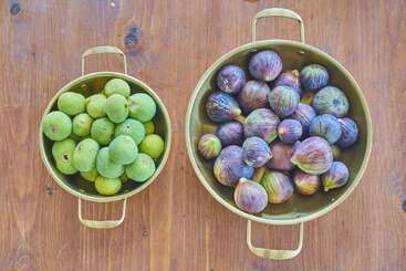 Two brass bowls sit on a wooden table. The left bowl contains green figs, while the right is filled with purple figs. Both bowls have handles.