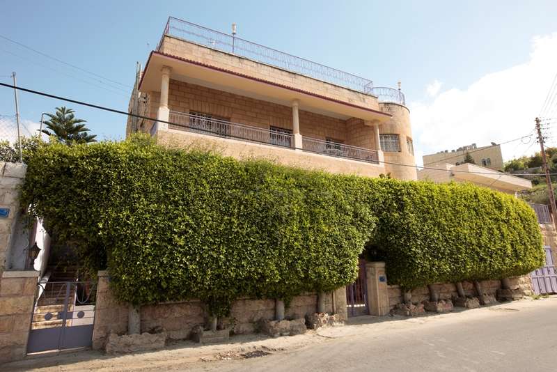 A imagem mostra uma casa grande com um quintal cercado, com um prédio de dois andares com um telhado plano e uma grande árvore ao fundo, cercada por uma área gramada.
