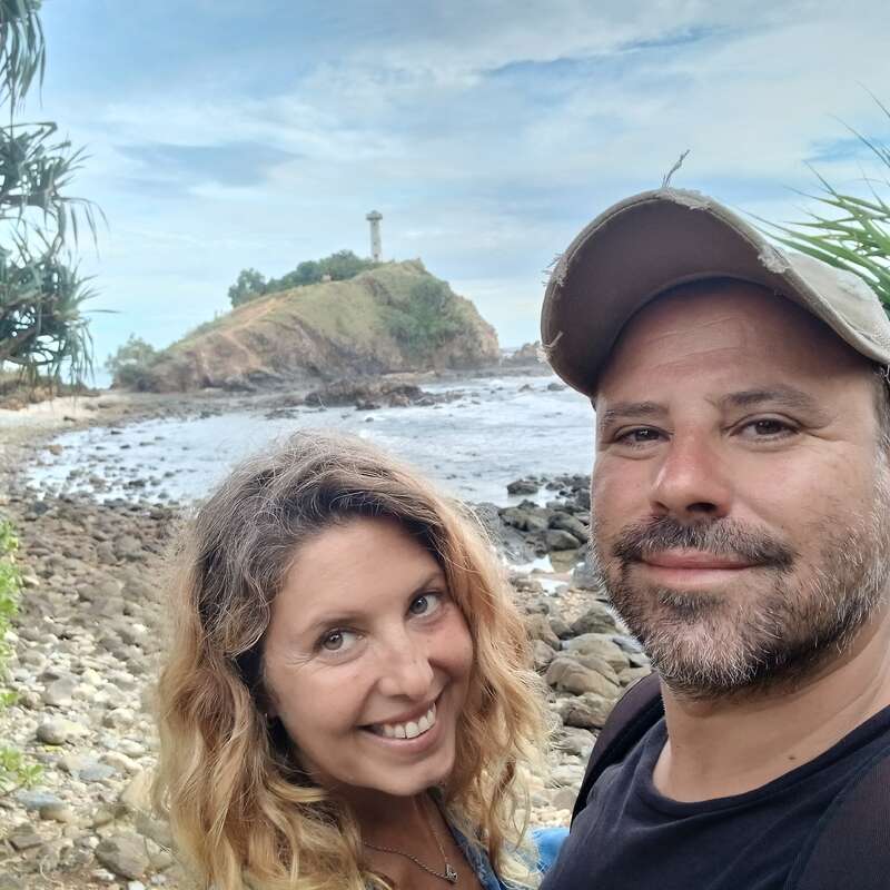 A smiling couple takes a selfie on a rocky beach with the ocean behind them. A small hill with a lighthouse sits in the background.