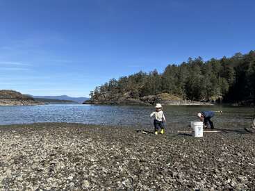 Uma criança com botas amarelas e um adulto estão numa costa rochosa à beira-mar, rodeados por colinas arborizadas, sob um céu azul e limpo.