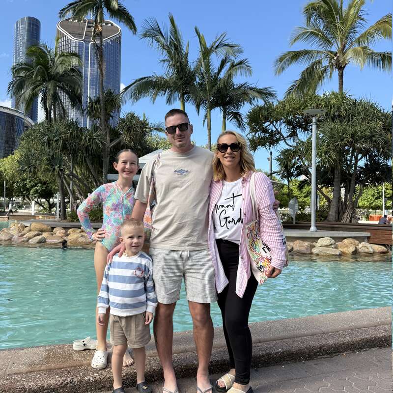 Una familia de cuatro miembros posa junto a una piscina, rodeada de palmeras y altos edificios modernos. Parecen felices, disfrutando juntos de un día soleado al aire libre.