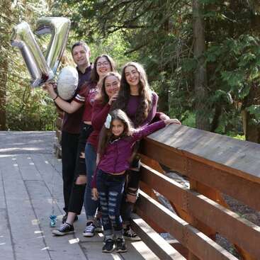 The image depicts a family of five posing on a bridge, with the father holding silver balloons in the shape of a \"V\" and a white volleyball.