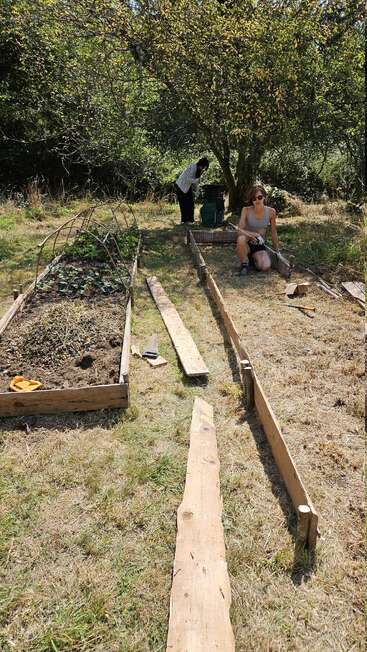 Deux personnes travaillent dans un jardin ensoleillé, entouré d'arbres. L'une est assise en train d'assembler un lit surélevé en bois, tandis que l'autre se tient près d'outils à l'ombre de l'arbre.