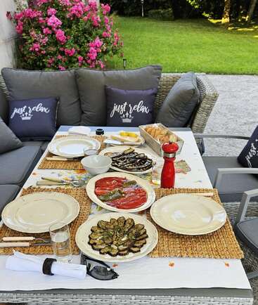Une salle à manger confortable en plein air est composée de coussins gris, de coussins "relax", de pain frais, de légumes grillés et d'assiettes invitantes, nichés à côté de fleurs roses colorées et d'une pelouse verte.