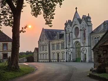 Un magnifique bâtiment historique européen se dresse sous un coucher de soleil radieux. Des branches d'arbres encadrent la scène, projetant des ombres sur la rue et la cour silencieuses et vides.
