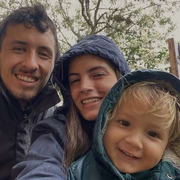 Une famille heureuse de trois personnes, portant des vestes à capuchon, sourit chaleureusement à l'appareil photo en plein air. L'arrière-plan montre des arbres, suggérant un environnement frais et naturel.