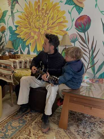 Ein Mann spielt Gitarre, während ein kleiner Junge zuhört. Sie sitzen drinnen auf einem gemusterten Teppich, umgeben von farbenfroher, geblümter Wandkunst und warmer Wohnkultur.