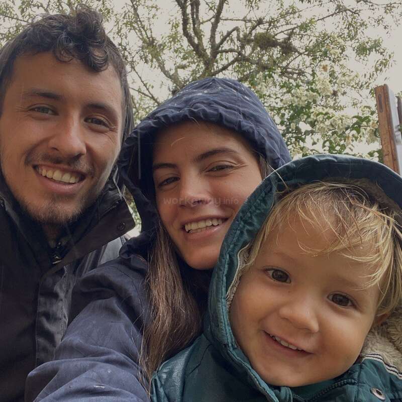 Eine glückliche dreiköpfige Familie mit Kapuzenjacken lächelt warm in die Kamera im Freien. Im Hintergrund sind Bäume zu sehen, die auf eine kühle, naturbelassene Umgebung hindeuten.
