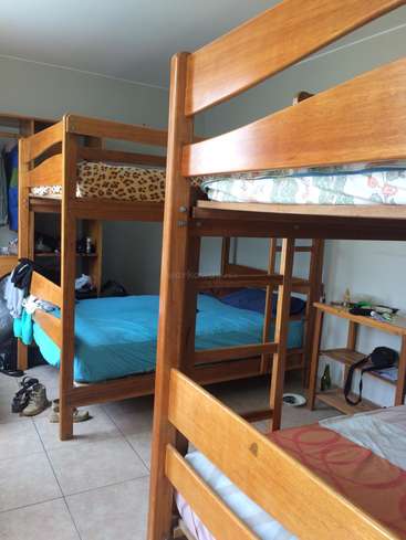 The image depicts a room with two wooden bunk beds, each with a blue sheet and a shelf at the foot, surrounded by various items on the floor and shelves.