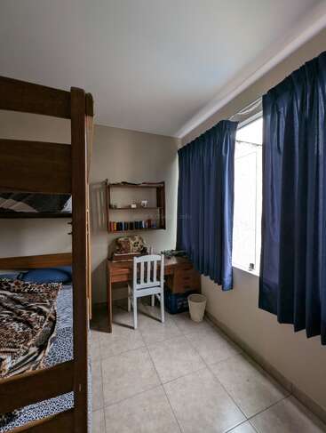 A cozy, tidy bedroom features bunk beds with patterned blankets, a wooden desk and chair, shelves with books, blue curtains, tiled floor, and a wastebasket.