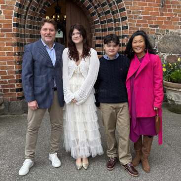 Four people stand smiling in front of a brick building. Two adults and two teenagers, all dressed formally, with flowers and an arched doorway visible behind them.