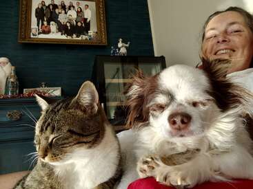 A smiling woman relaxes with her eyes closed cat and fluffy dog. Family photos and home decor create a cozy, warm, and inviting atmosphere in the background.