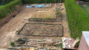 This image shows a neatly organized garden with several rectangular beds, some covered with soil or mulch, surrounded by green hedges, and various gardening tools and materials.