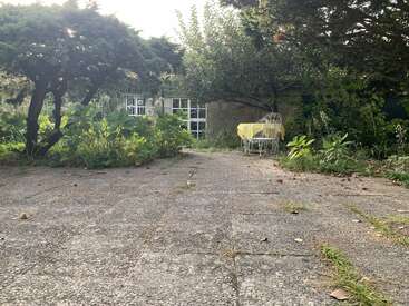 Une scène de jardin paisible avec de la verdure envahissante, des pavés en béton, des chaises en fer et une table recouverte d'un tissu jaune à l'ombre de grands arbres.