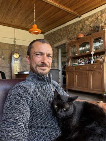 Ein Mann in einem gemusterten Pullover sitzt drinnen und lächelt, während eine schwarze Katze auf seinem Schoß liegt. Eine Holzdecke, Steinwände und Vintage-Möbel sorgen für eine gemütliche Atmosphäre.