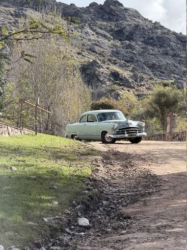 Ein hellblaues Oldtimer-Auto steht auf einem Feldweg in der Nähe einer Wiese, im malerischen Hintergrund sind felsige Hügel und vereinzelte Bäume zu sehen.
